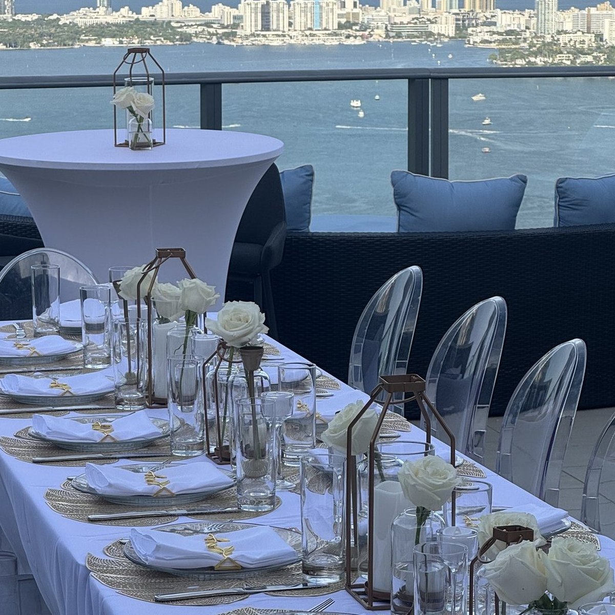A long white-linen table set for a corporate dinner on a Miami bayfront terrace, with ghost chairs, white roses and the skyline across the water