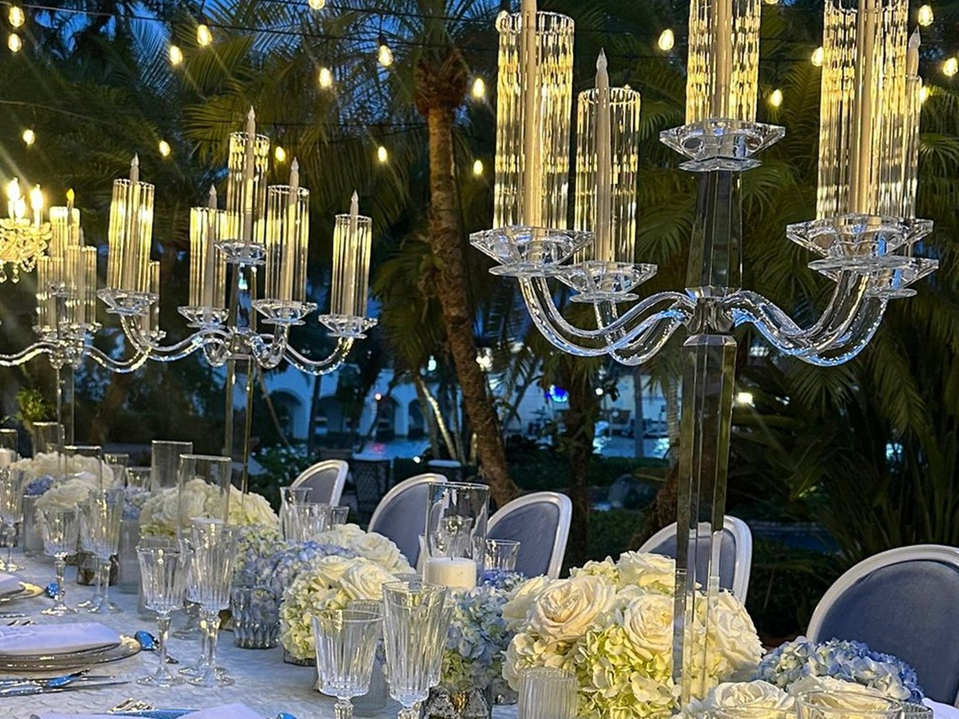 A resident dinner on a Miami pool deck at dusk, crystal candelabra and white florals under festoon lights