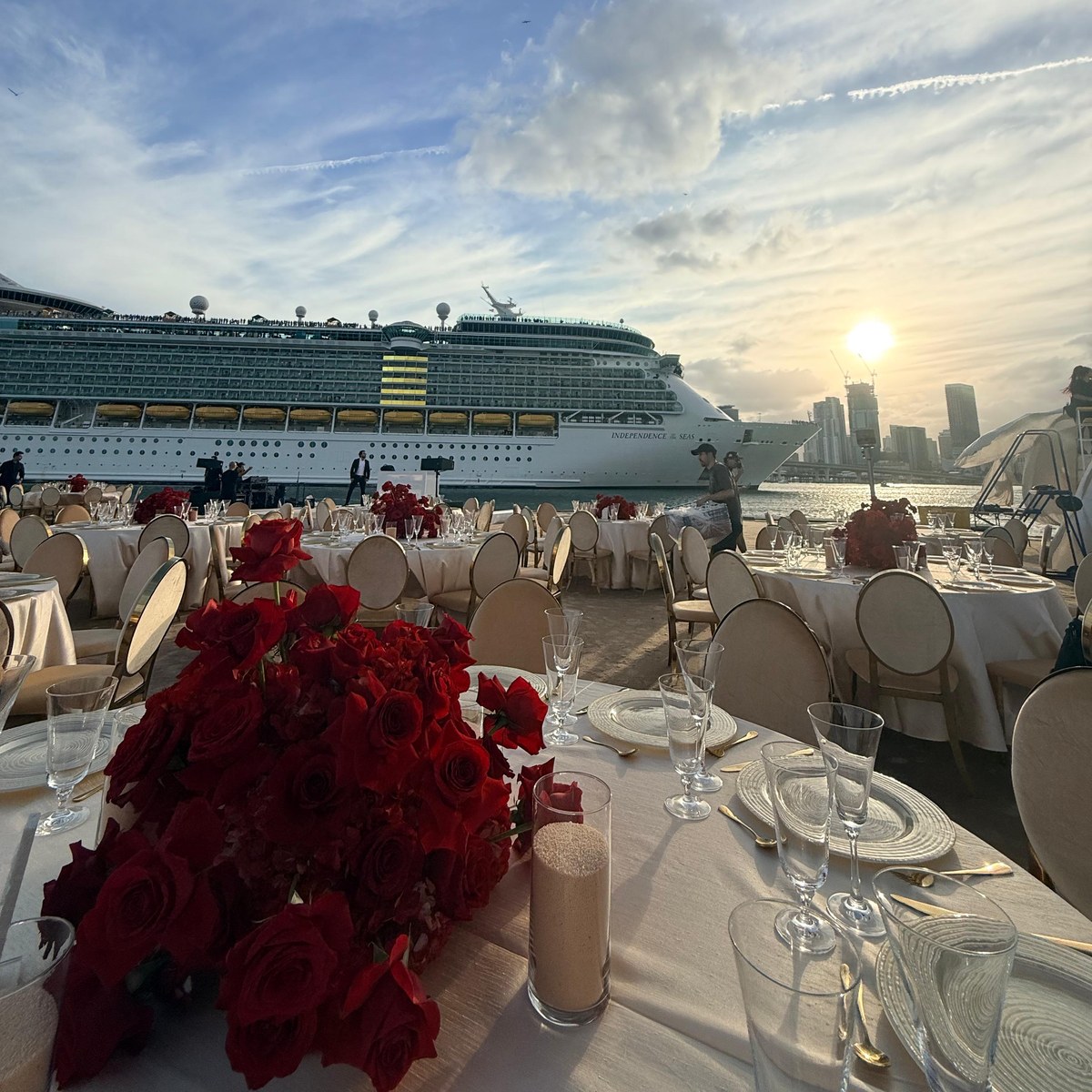 A waterfront reception at sunset in Miami: round tables in cream linen with red rose centrepieces and gold cutlery, the bay and the skyline behind