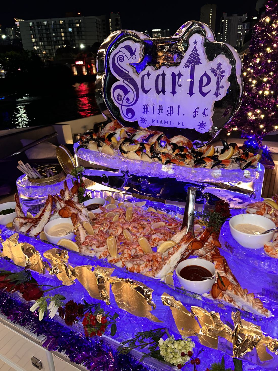 A tiered ice-carved raw bar lit from within: lobster tails, shrimp and stone crab claws with lemon, gold leaf and sauces, on a Miami waterfront terrace at night